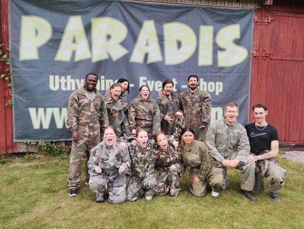 Ett kompisgäng på 12 personer som poserar framför Paradis Paintball & Äventyrs lada och ska snart spela paintball.