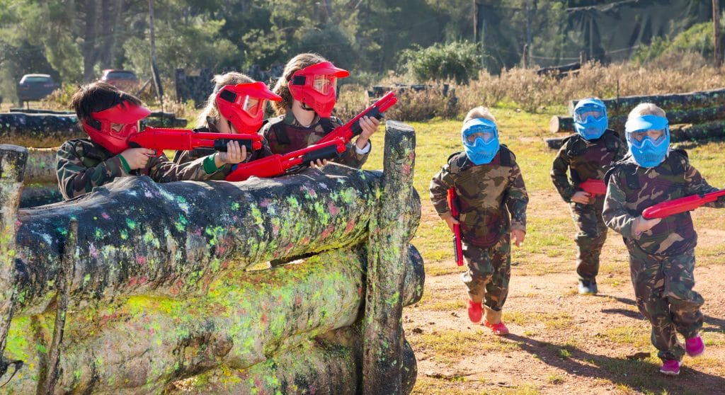 Två motståndarlag med glada barn som spelar på en paintballbana utomhus.
